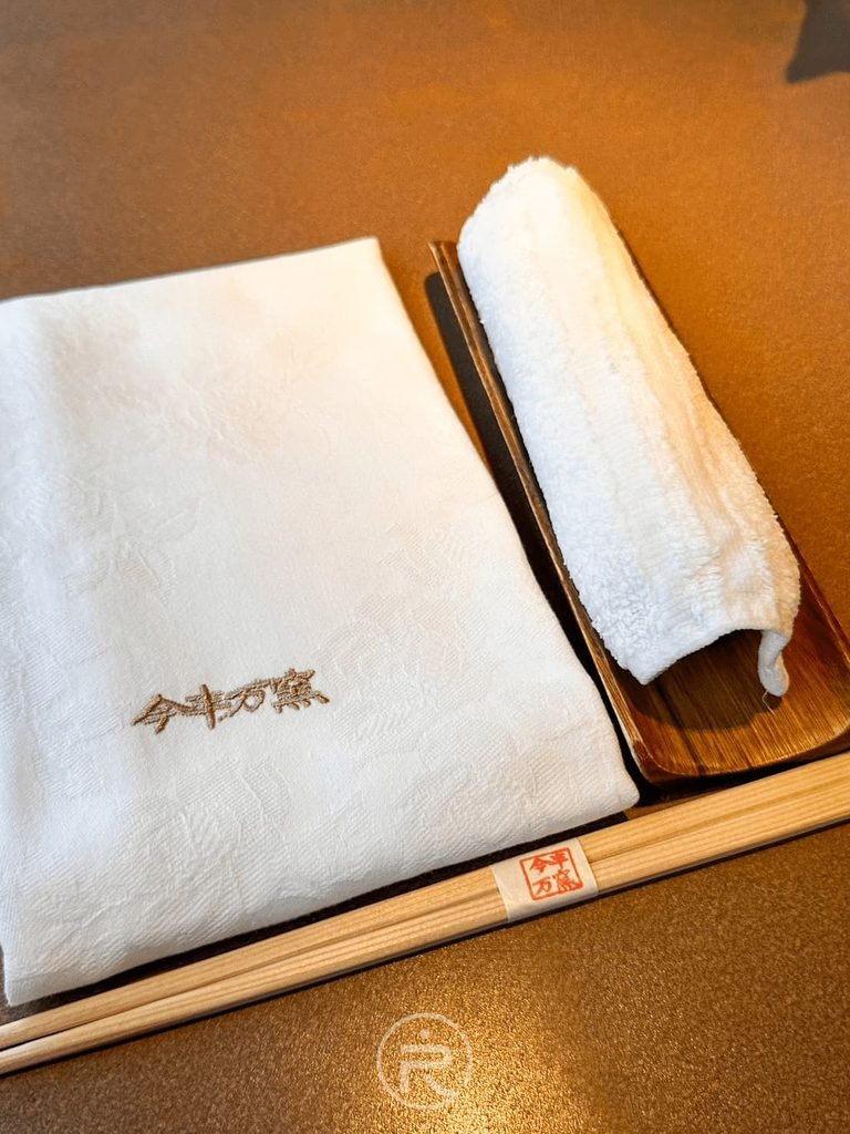 [餐具擺設]： 繡有「今半萬窯」字樣的白色圍兜餐巾、日式木筷與熱毛巾擺設。