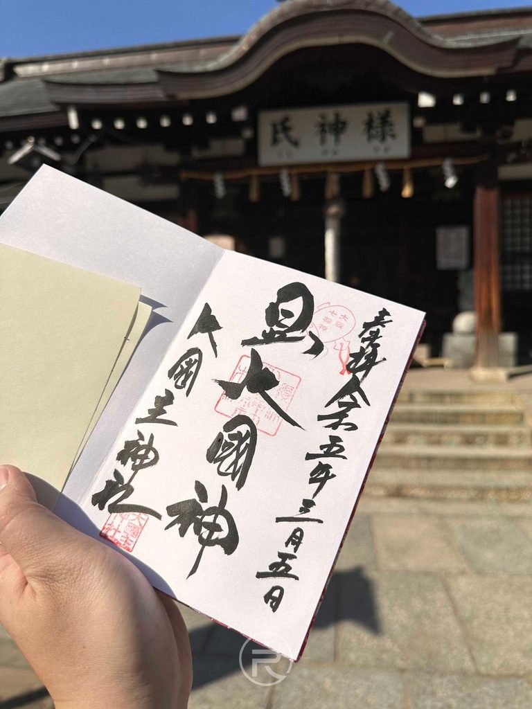 [手持御朱印與本殿]： 大阪大國主神社手寫御朱印實拍，上書「大國神」墨寶與氏神樣拜殿合影。
