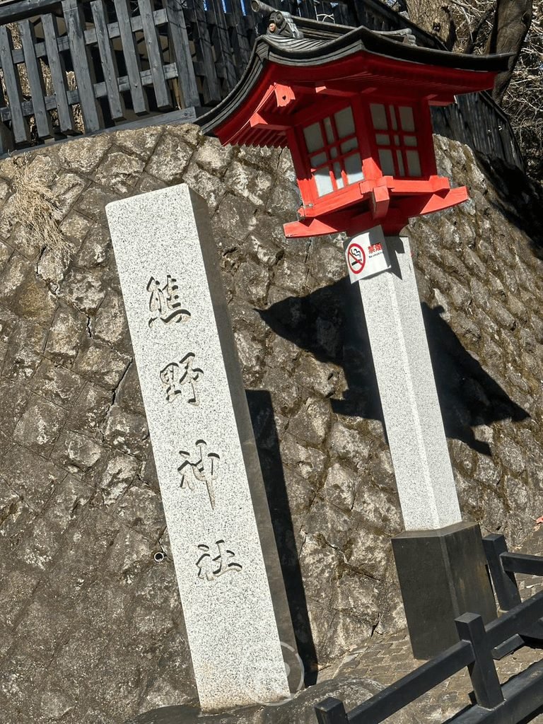  [社號石碑與朱紅燈籠]： 新宿十二社熊野神社入口石碑與石造朱紅宮燈，在樹影下顯得古樸莊嚴。