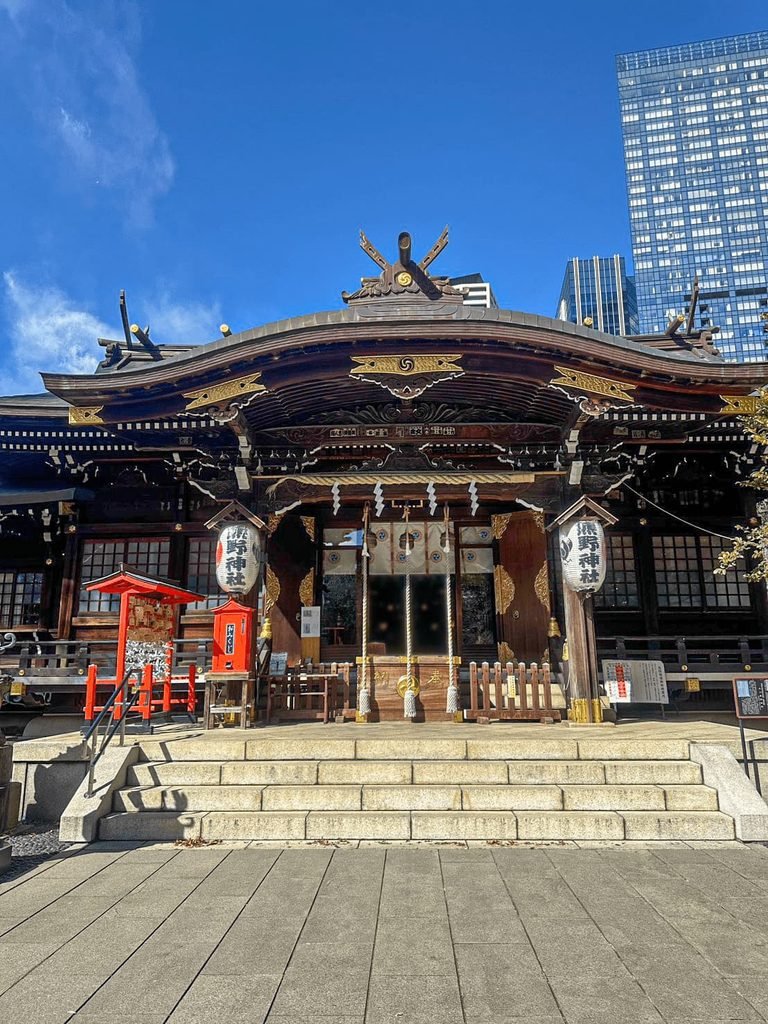 [本殿與高樓同框]： 熊野神社本殿正面全景，後方現代摩天高樓林立形成強烈古今對比。