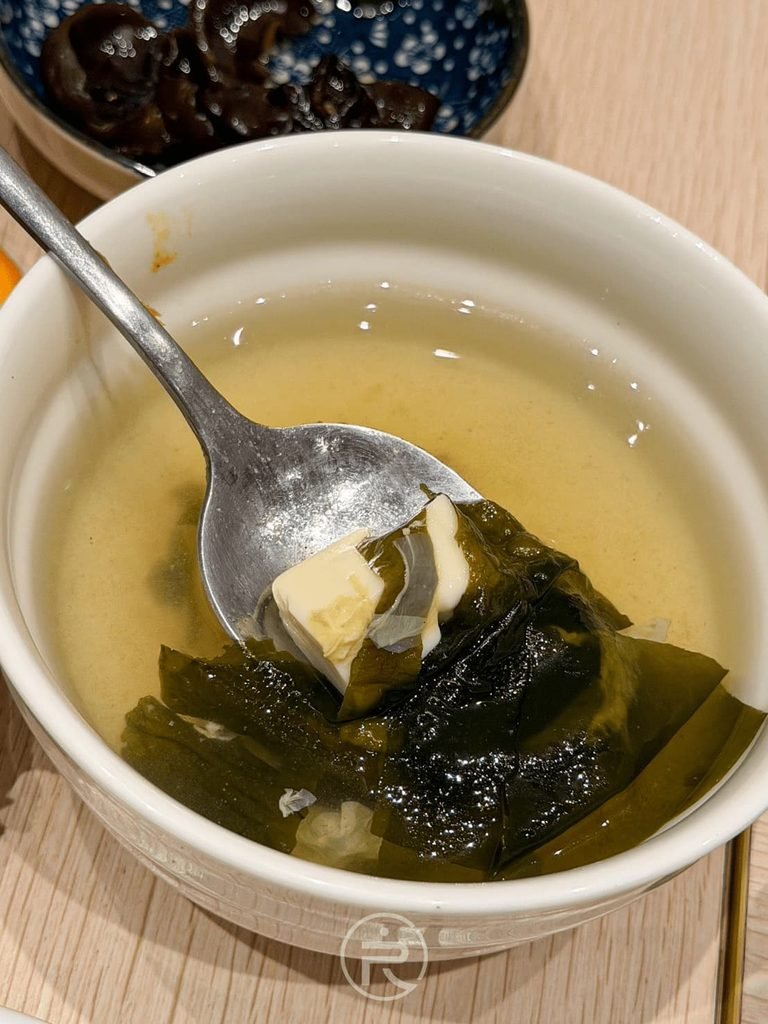  [湯匙舀嫩豆腐]： 湯匙撈起海帶芽豆腐湯中的大塊嫩豆腐與海帶芽。