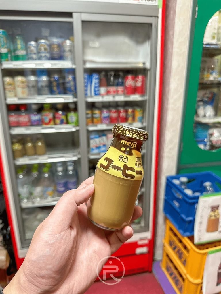  [手拿咖啡牛奶]： 在飲料冷藏櫃前手持一瓶剛買的冰鎮「明治咖啡牛奶（Meiji Coffee）」。