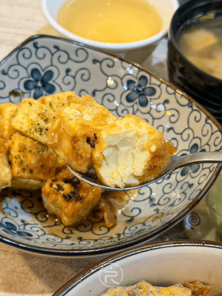  [舀起揚出豆腐]： 湯匙舀起一顆切開的現炸揚出豆腐，外皮薄酥內裡軟嫩。
