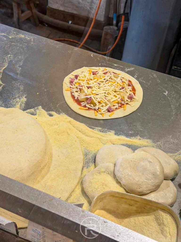 [進爐前鋪料披薩]： 剛鋪滿新鮮起司與豐富食材、準備送入高溫柴燒爐烘烤的披薩餅皮。