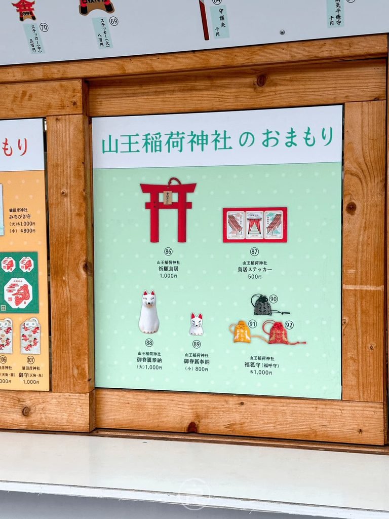 [山王稻荷御守看板]： 木架上展示的山王稻荷神社專屬白狐御守與開運祈福說明。
