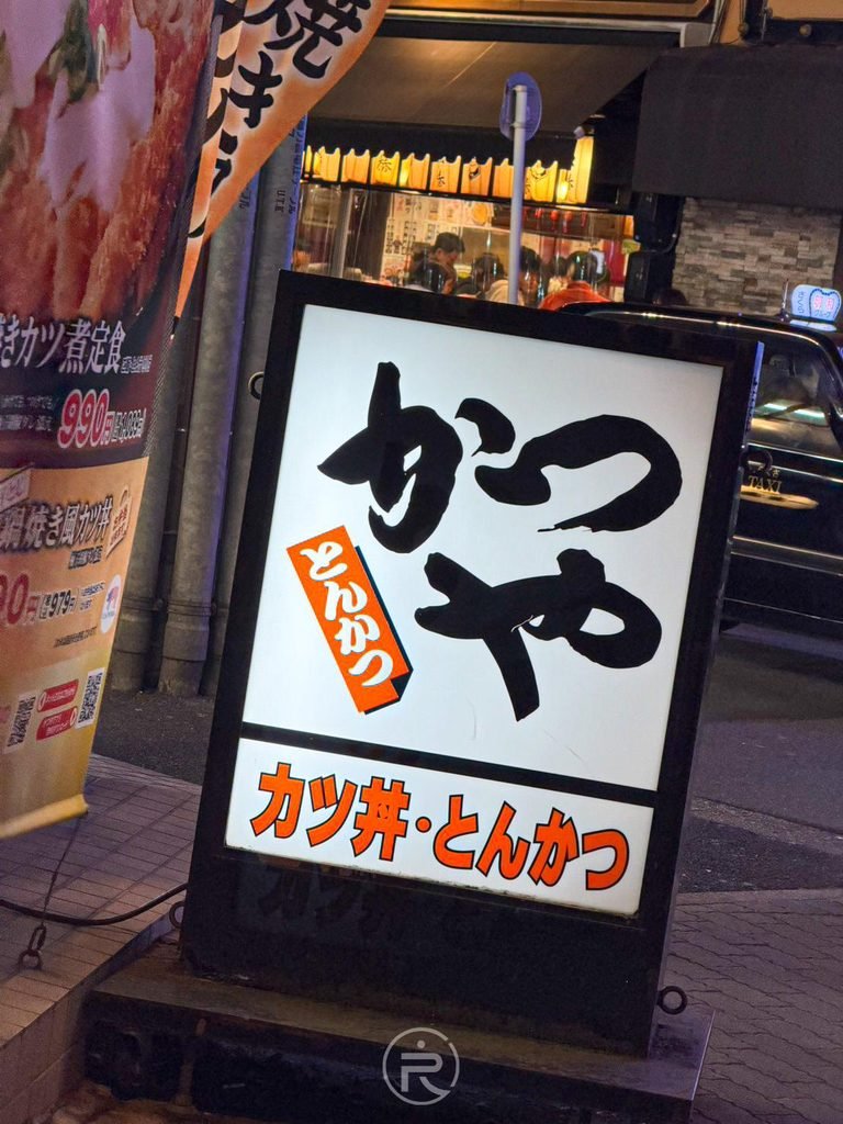  [人行道發光立牌]： 人行道旁醒目的黑白字「かつや カツ丼・とんかつ」夜間發光廣告燈箱。