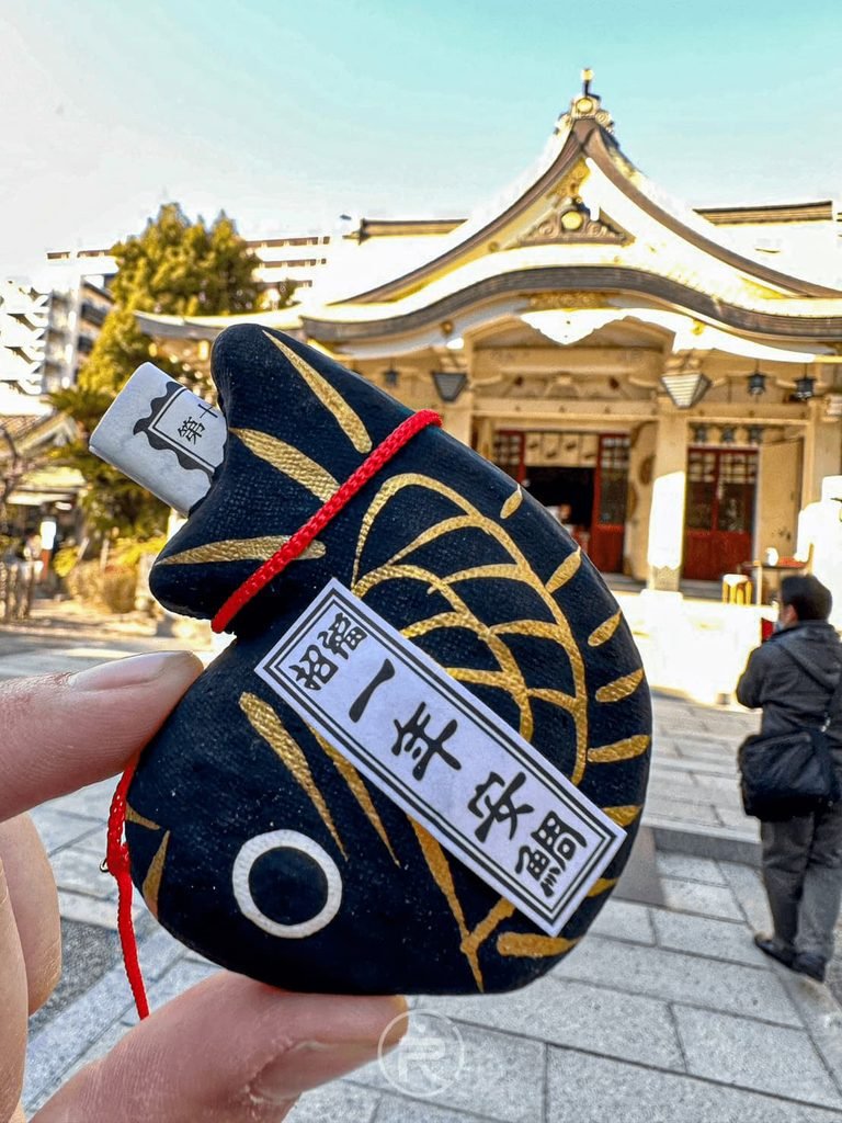 [手持一年安鯛神籤]： 手持黑色的「一年安鯛（鯛魚籤）」祈福御守，背景為陽光下的神社本殿。