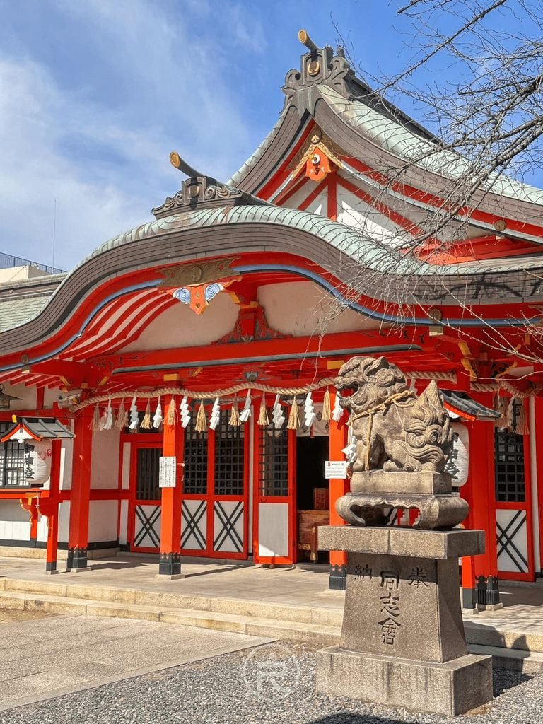 [朱紅拜殿正面]： 晴空下的玉造稻荷神社御本殿，鮮豔朱紅梁柱、白牆與石造狛犬守護神獸。