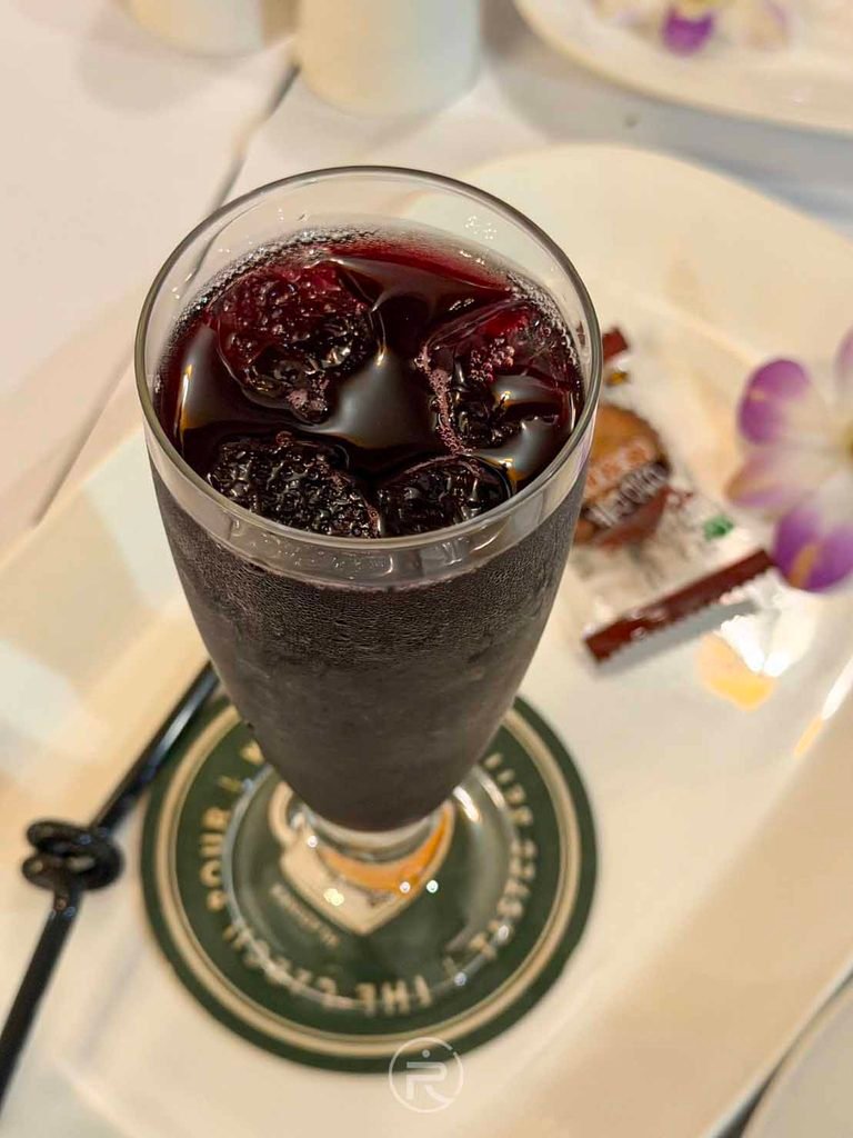  [冰涼葡萄汁全杯]： 盛裝在典雅高腳玻璃杯中的深紫色純葡萄汁，搭配蘭花裝飾。