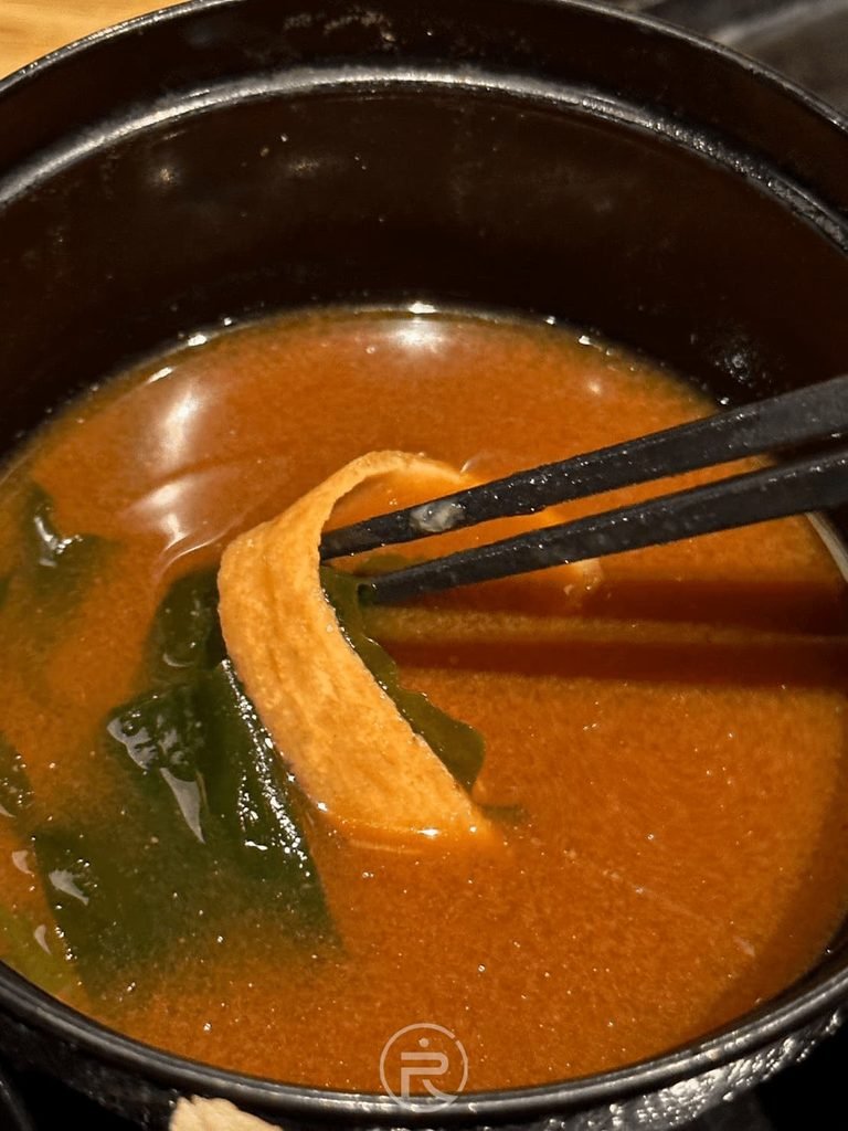  [紅味噌湯配料]： 筷子舀起套餐附贈的日式紅味噌湯，內有滑嫩海帶芽與吸汁豆皮。