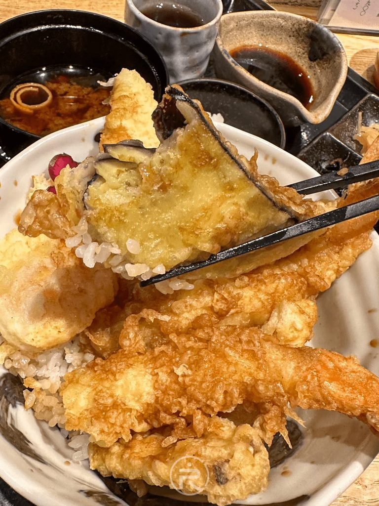  [夾起野菜天婦羅]： 筷子夾起香脆野菜天婦羅配料，色澤金黃誘人。